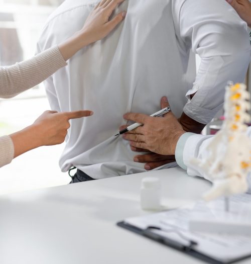 Doctor explains back pain to patient in hospital office, pointing with pen. Patient listens attentively, seeking relief. Anatomical model aids comprehension